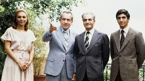 Four adults in formal attire stand outdoors against a backdrop of green foliage and a railing — a public domain photograph from The Shah of Iran archive documenting the Pahlavi era.