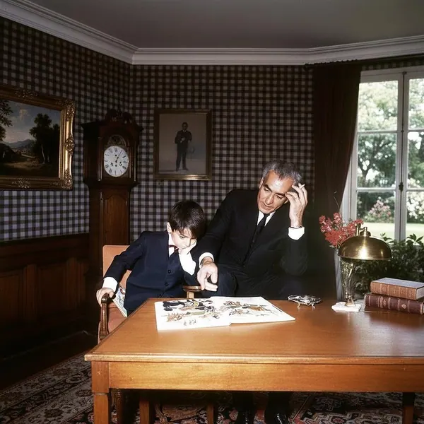His Majesty, the Shah of Iran and a boy in dark suits look at a book on a wooden desk in a room — a public domain photograph from The Shah of Iran archive documenting the Pahlavi era.