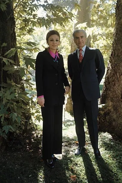 His Majesty, the Shah of Iran and woman in dark formal suits stand side by side outdoors among large trees — a public domain photograph from The Shah of Iran archive documenting the Pahlavi era.