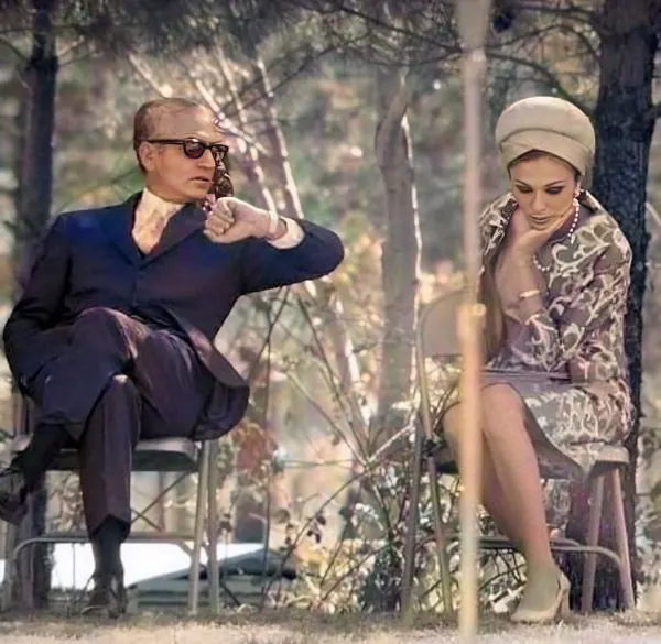 His Majesty, the Shah of Iran in a dark suit and Farah Pahlavi, Empress of Iran, in a patterned dress sit outdoors among trees — a public domain photograph from The Shah of Iran archive documenting the Pahlavi era.