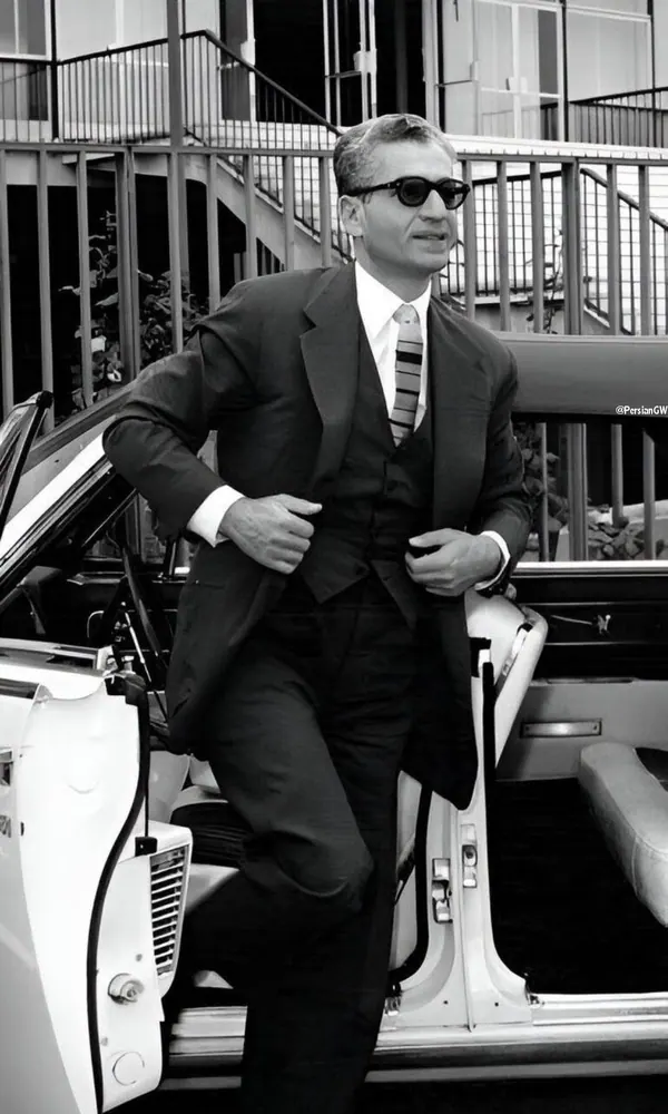 His Majesty, the Shah of Iran in a dark three-piece suit and sunglasses leans against an open white car door — a public domain photograph from The Shah of Iran archive documenting the Pahlavi era.