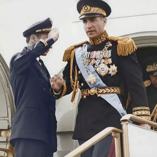 His Majesty, the Shah of Iran in a decorated military uniform stands on stairs while another uniformed man salutes him — a public domain photograph from The Shah of Iran archive documenting the Pahlavi era.