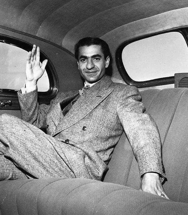 His Majesty, the Shah of Iran in a double-breasted suit sits in a car backseat and waves with one hand — a public domain photograph from The Shah of Iran archive documenting the Pahlavi era.
