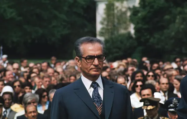His Majesty, the Shah of Iran in a suit and glasses stands in front of a large, blurred crowd of people — a public domain photograph from The Shah of Iran archive documenting the Pahlavi era.