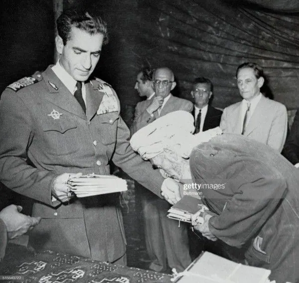 His Majesty, the Shah of Iran in military uniform hands a stack of papers to a person bowing forward in a coat — a public domain photograph from The Shah of Iran archive documenting the Pahlavi era.
