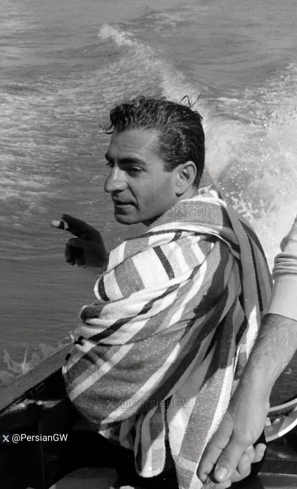 His Majesty, the Shah of Iran with a striped towel draped over his shoulders sits on a boat amidst splashing water — a public domain photograph from The Shah of Iran archive documenting the Pahlavi era.