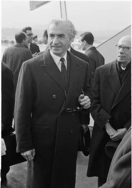 His Majesty, the Shah of Iran in a dark overcoat and tie stands outdoors among other men in formal attire — a public domain photograph from The Shah of Iran archive documenting the Pahlavi era.