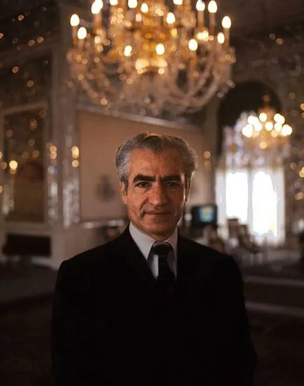 His Majesty, the Shah of Iran in a dark suit and tie stands before a large, ornate crystal chandelier — a public domain photograph from The Shah of Iran archive documenting the Pahlavi era.