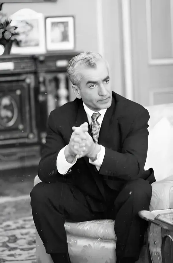His Majesty, the Shah of Iran in a dark suit and tie sits in a chair with hands clasped together — a public domain photograph from The Shah of Iran archive documenting the Pahlavi era.