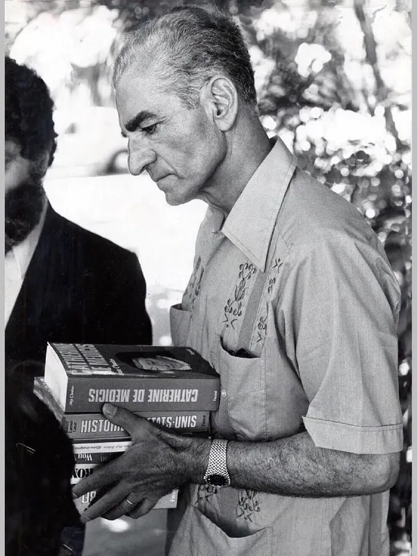 An older man in a short-sleeved shirt holds a stack of books while standing outdoors — a public domain photograph from The Shah of Iran archive documenting the Pahlavi era.