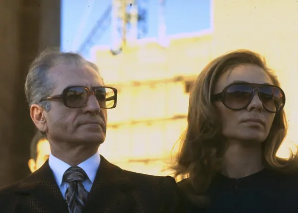 An older man in a suit and Farah Pahlavi, Empress of Iran, with long hair both wear dark sunglasses — a public domain photograph from The Shah of Iran archive documenting the Pahlavi era.