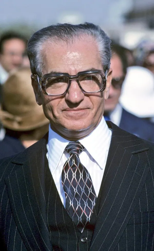An older man with glasses wears a pinstripe suit and patterned tie in a close-up portrait — a public domain photograph from The Shah of Iran archive documenting the Pahlavi era.