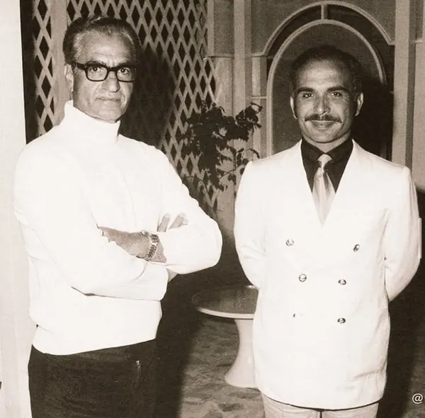 Two middle-aged men stand side by side, one in a white turtleneck and the other in a white suit — a public domain photograph from The Shah of Iran archive documenting the Pahlavi era.